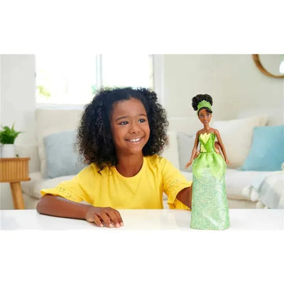 A smiling girl in a yellow shirt holds a Mattel Disney Princess Core Doll of Tiana, dressed in a green gown, on a table.