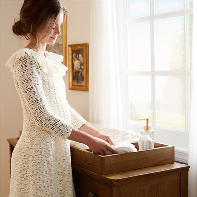 A woman in a white dress arranges linens in a wooden changing table with a quilted topper and decorative items nearby.