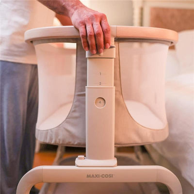 Maxi-Cosi Iora Baby Bassinet Bedside Sleeper in Truffle with adjustable height and mesh sides, being adjusted by a hand.