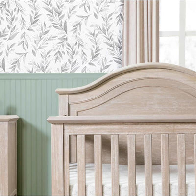 Rustic 4-in-1 Convertible Curve Top Crib in Sandbar finish, showcased in a nursery with leaf-patterned wallpaper and green paneling.