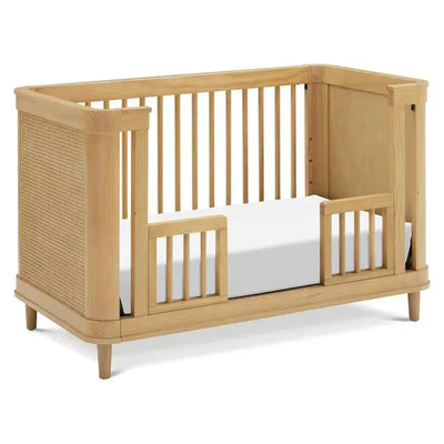 3-in-1 convertible crib in honey finish featuring a cane side, open slats, and a white mattress.