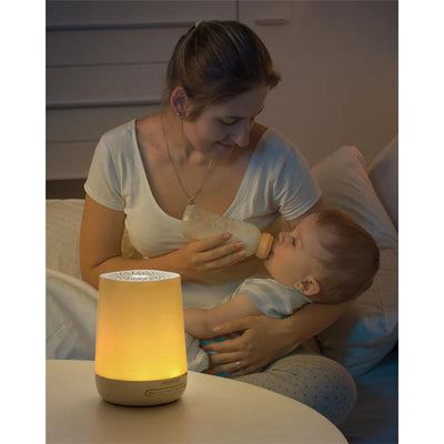 A mother bottle-feeds her baby at night, with a glowing Momcozy white noise machine on a table nearby.