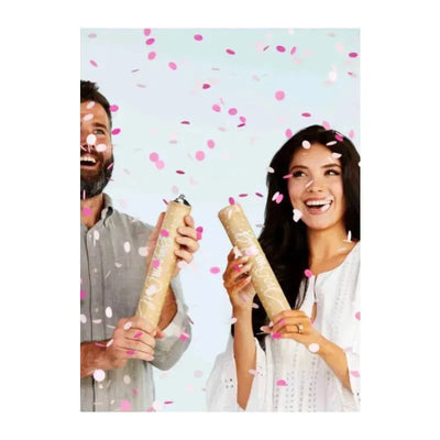 A man and a woman joyfully hold pink confetti cannons, surrounded by pink confetti in celebration of a gender reveal.