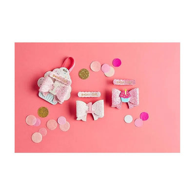 White birthday hair clips from Mud Pie, featuring a cupcake design and sparkly bows, arranged on a pink background with confetti.