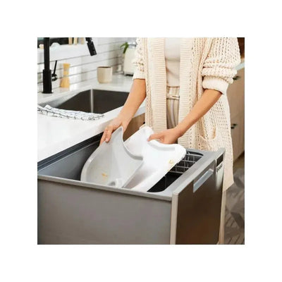 A woman placing a removable, white tray from the Munchkin 360° Cloud Baby High Chair into a dishwasher.