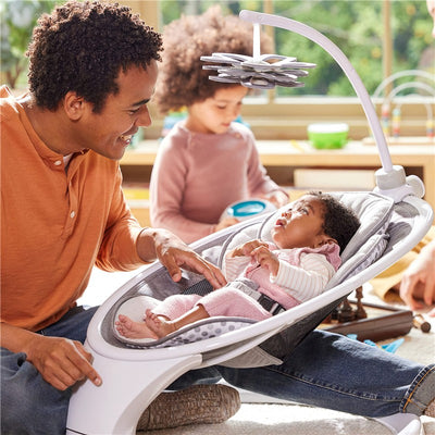 High-contrast Munchkin Baby Bloom mobile hangs above a baby in a bouncer, with a father engaging and a sibling playing nearby.