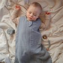 Organic cotton muslin baby wearable blanket in Tradewinds, with a sleeping baby and soft toys on a white blanket.