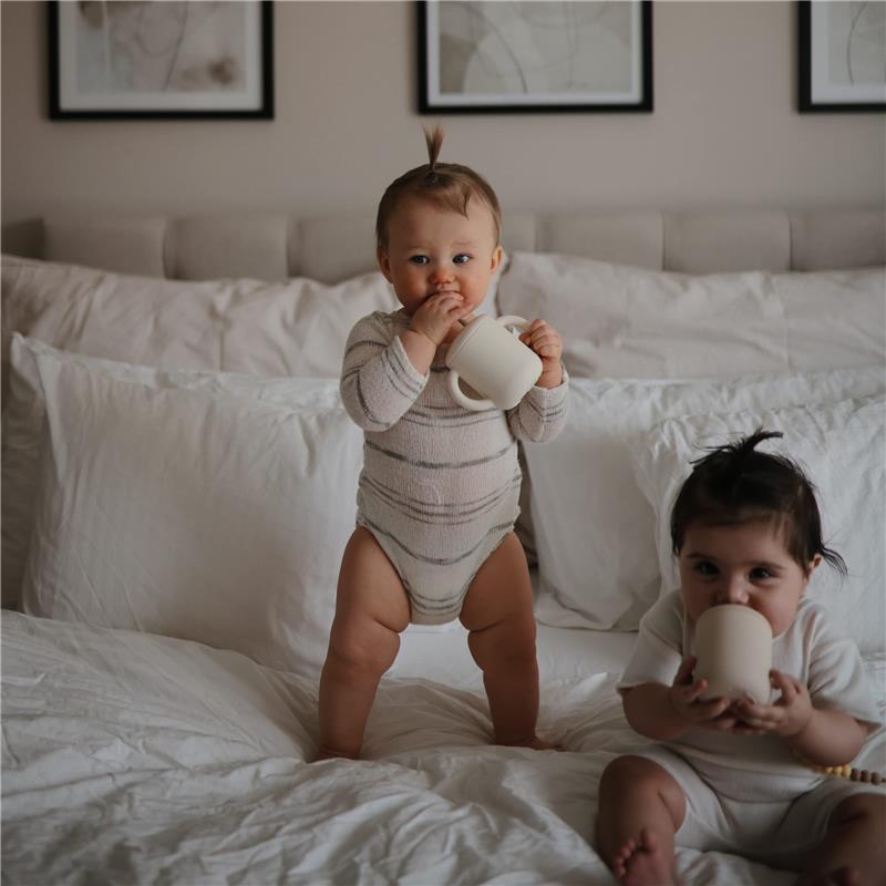 Two toddlers are sitting on a bed, one standing while holding a lilac silicone training cup with straw.