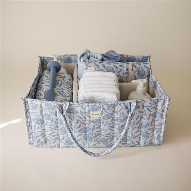 Mushie Baby Diaper Caddy with blue flower design, containing diapers, wipes, a bottle, and a soft blanket.