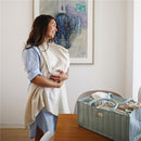 A woman in a light blue dress smiles while holding a baby blanket and standing next to a storage caddy for newborn essentials.