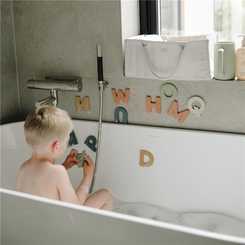 A young child sits in a bubble-filled bathtub, playing with bath letters while a white Mushie Baby Diaper Caddy is visible nearby.