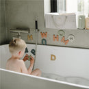 A young child sits in a bubble-filled bathtub, playing with bath letters while a white Mushie Baby Diaper Caddy is visible nearby.