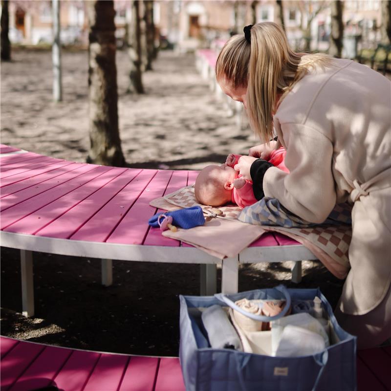A caregiver adjusts a diaper on a baby lying on a pink table in a park, with a blue diaper caddy nearby.
