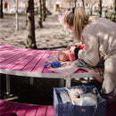 A caregiver adjusts a diaper on a baby lying on a pink table in a park, with a blue diaper caddy nearby.