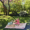 A young girl in pink pajamas sits on a picnic blanket in a sunny garden, playing with a toy camera, surrounded by foliage and flowers.