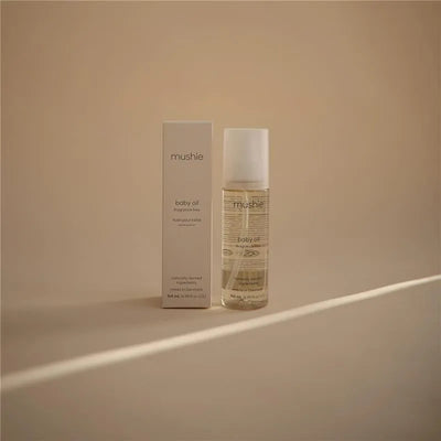 Mushie baby oil, 145 ml, in a transparent bottle next to its white box, both displayed against a soft beige background.