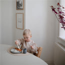 Toddler wearing a long sleeve water-resistant bib, eating from a plate with rice and food, at a light-filled dining area.