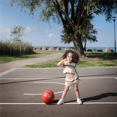 A young child with curly hair stands on a basketball court, striking a pose next to a red basketball.