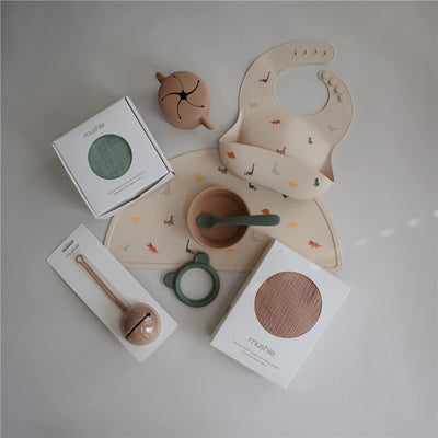 Silicone baby bib featuring a dinosaur design, accompanied by feeding accessories from Mushie, including a bowl and spoon.