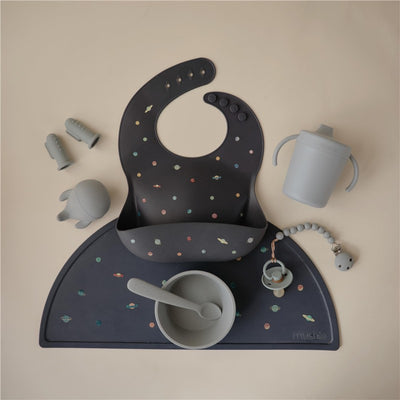 Mushie silicone baby bib with a planet design, accompanied by a gray bowl, spoon, cup, and silicone toys.