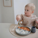 A toddler with short hair uses a light blue silicone spoon to eat from a divided gray plate with food.
