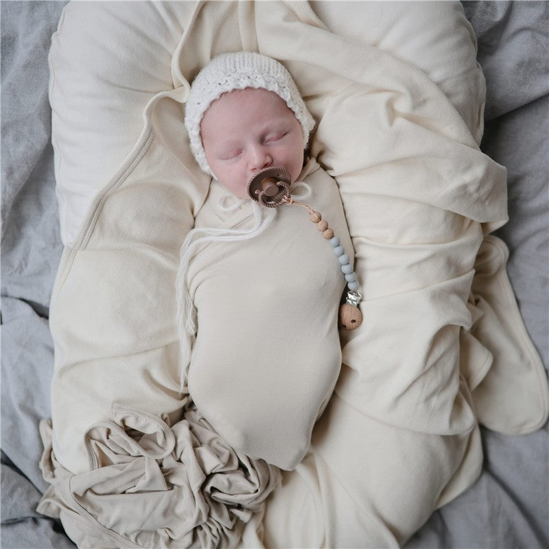 Mushie Eva Pacifier Clip in Stone attached to a sleeping baby swaddled in soft, neutral-toned fabrics.