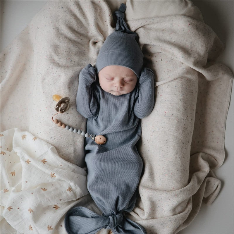 Mushie Eva Pacifier Clip in stone color attached to a sleeping baby wrapped in a soft blanket, wearing a matching gray outfit.