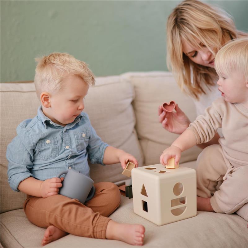 Children playing with a shape sorter toy on a couch; a boy holds a cup while another child places shapes into the toy.