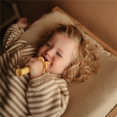 Toddler smiling while holding a yellow Mushie silicone flower training toothbrush, wearing a striped long-sleeve top.