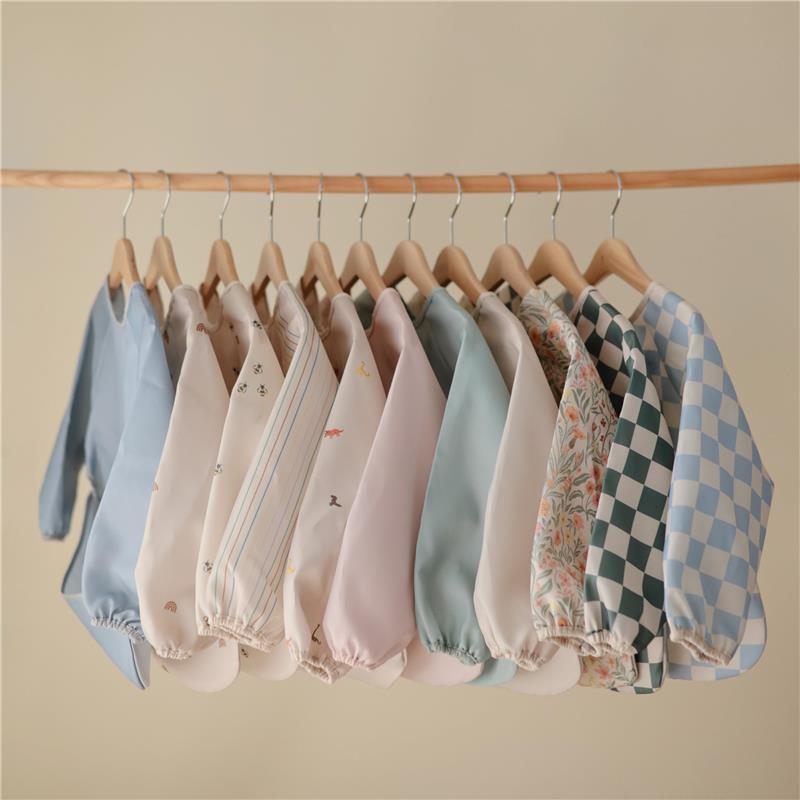 Water-resistant, adjustable long sleeve baby bibs in various colors and patterns hanging on a wooden rack.