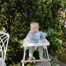 Baby wearing a blue check long sleeve bib in a high chair, eating, with a bowl and cup on the tray surrounded by greenery.