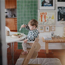 Baby in a high chair wearing a blue check long sleeve bib, eating from a bowl of food with a fork, in a styled kitchen.