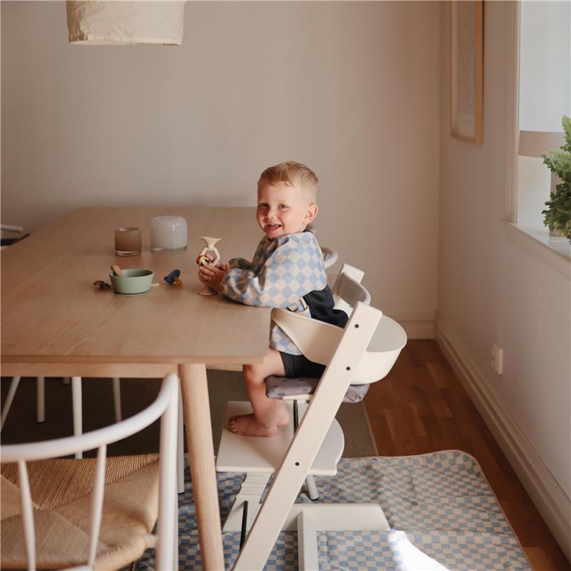 Baby wearing a blue check adjustable long sleeve bib sitting at a wooden table, smiling while holding a toy.