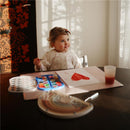 A young child wearing a bow and a long sleeve baby bib is seated at a table, holding a paintbrush and smiling, with a painted heart on paper and art supplies nearby.