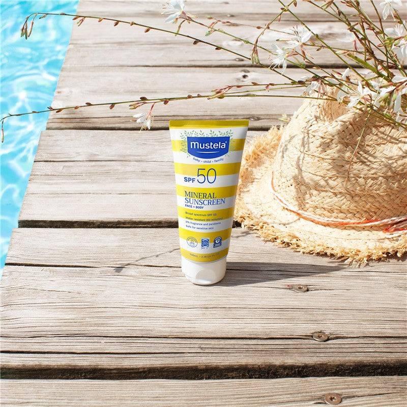 Mustela Mineral Sunscreen Lotion SPF 50, 3.38 fl oz, placed on wooden surface near a straw hat and pool water.