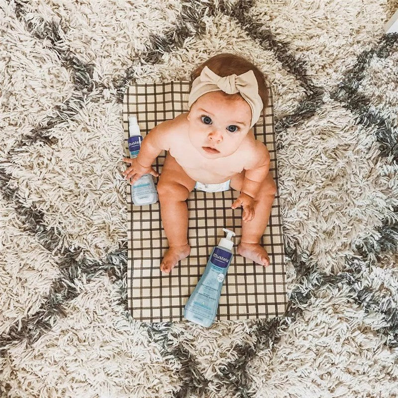 A baby sits on a checkered mat surrounded by two bottles of Mustela Hair Styler & Skin Freshener, featuring light blue packaging.