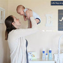 A mother joyfully lifts her baby in a nursery, with Mustela hair styler and skin freshener products visible on the shelf.