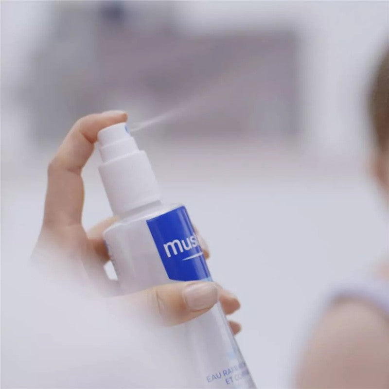 Mustela Hair Styler & Skin Freshener being sprayed from a pump bottle, held by a hand, with blurred background.