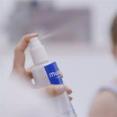 Mustela Hair Styler & Skin Freshener being sprayed from a pump bottle, held by a hand, with blurred background.