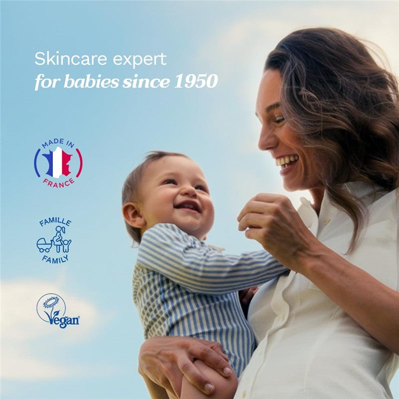 Mustela Hair Styler & Skin Freshener bottle beside a smiling mother holding her happy baby outdoors with blue sky.