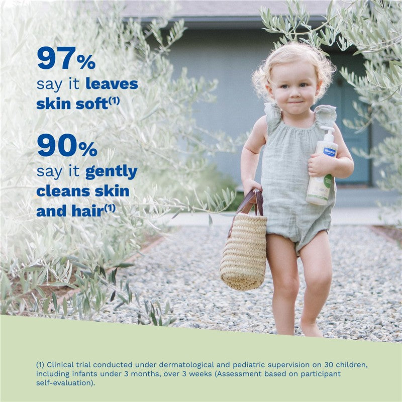 Mustela Organic Cleansing Gel bottle held by a smiling child in a light green romper, walking on a pebbled path surrounded by olive branches.