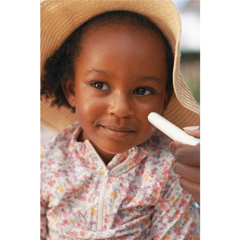 A young child with curly hair and a sunhat receives sunscreen applied to her face, featuring the Mustela SPF 50 Mineral Sunscreen Stick.