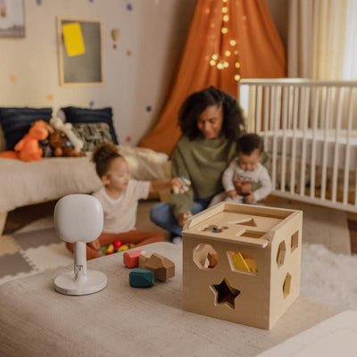 Nanit Pro Camera on a table in a child’s playroom, with a mother interacting with her two children and a colorful shape-sorting toy nearby.