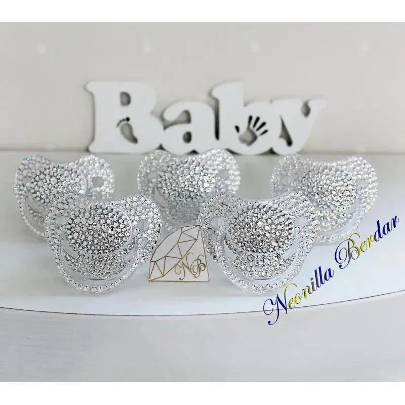 Crystal-embellished Neonilla orthodontic pacifiers displayed in a row on a white surface, with a decorative baby sign in the background.