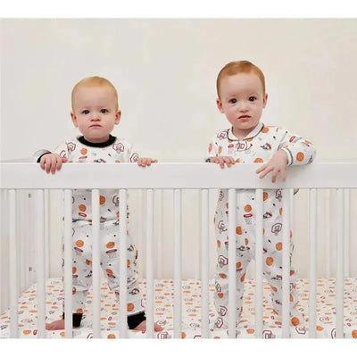 Two babies in a crib wearing Noomie baby basketball court zipper footies with basketball-themed designs.