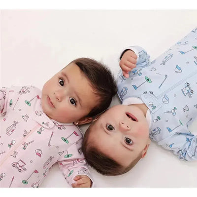 Two infants lie side by side on a white surface, one in a pink golf-themed footie with patterns of golf carts and flags, and the other in a light blue footie decorated with golf clubs and balls.