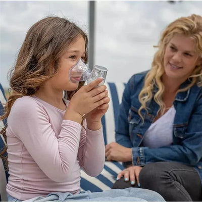A young girl uses a nebulizer with a mask, while a smiling woman observes her attentively.
