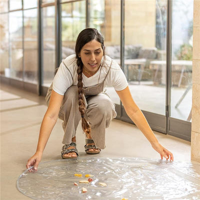 A woman kneels on a floor, placing a transparent Nuby waterproof floor mat down, with small food pieces scattered on it.