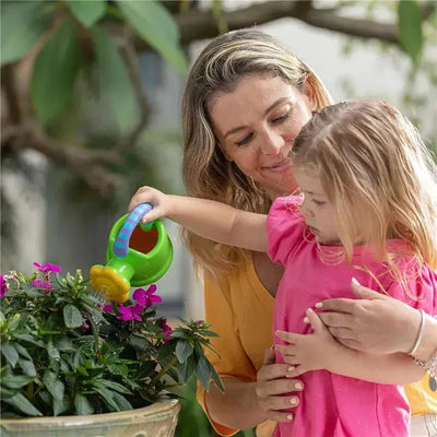 A woman helps a child use a green watering can bath toy to water pink flowers in a planter.