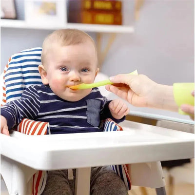 Green baby feeding spoon being used to feed a smiling baby in a high chair.